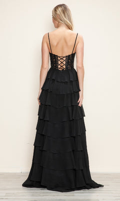 A woman stands with her back to the camera, wearing the Poly USA 9698 Long Tiered Corset Prom Dress—floor-length, black, a-line with a corset-style bodice—against a plain light-colored wall.