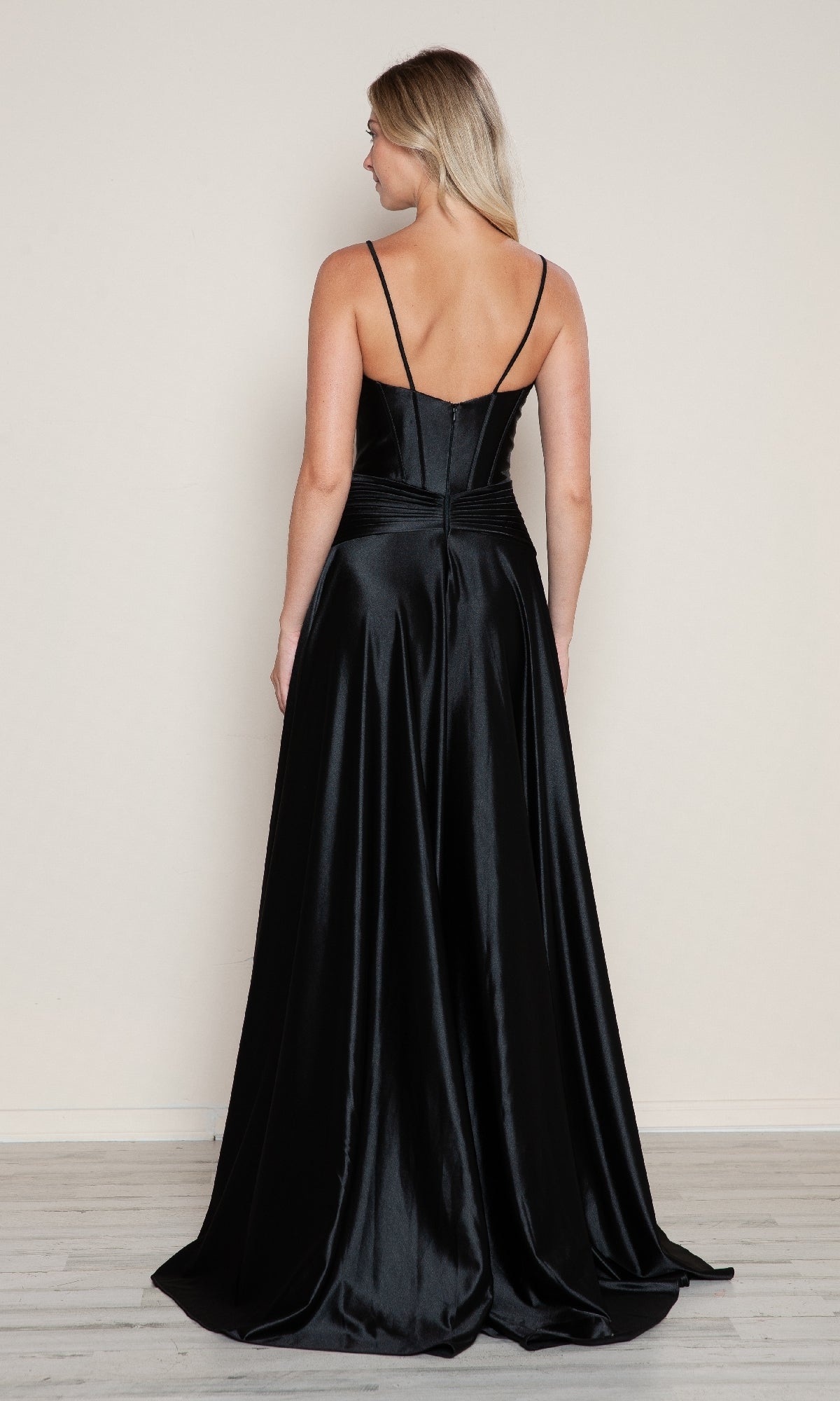 A woman faces away, wearing the Cut-Out Long A-Line Prom Dress Poly USA 9700—an elegant black satin gown with thin straps and an A-line silhouette—against a plain, light background.
