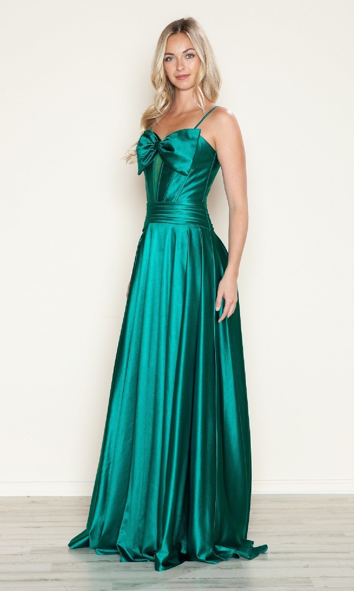 A woman stands against a plain wall wearing the Bow-Front Long Corset Prom Dress: Poly USA 9702—an emerald green, floor-length satin gown with thin straps, a bow accent on the bodice, and an elegant cut-out corset design.