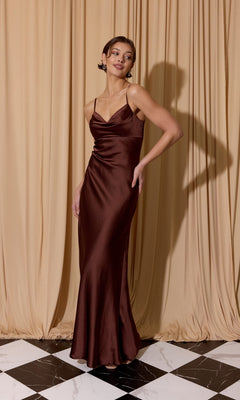 A woman in the Eureka 9711 Cowl-Neck Long Satin Prom Dress in brown stands on a black and white tiled floor before beige curtains.