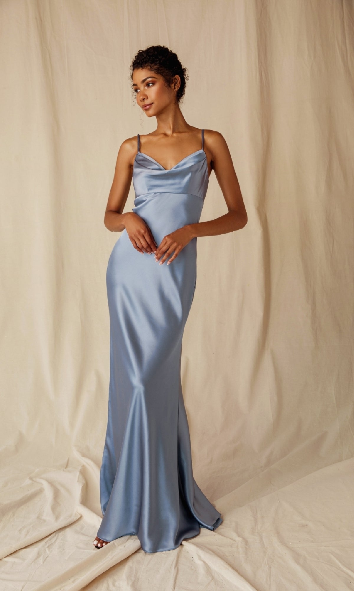 A woman in the Eureka 9711 Cowl-Neck Long Satin Prom Dress with spaghetti straps, light blue in color, stands before a cream-colored fabric backdrop.
