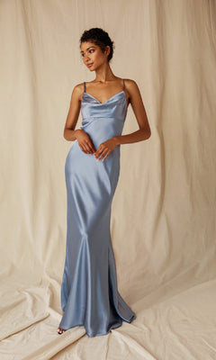 A woman in the Eureka 9711 Cowl-Neck Long Satin Prom Dress with spaghetti straps, light blue in color, stands before a cream-colored fabric backdrop.
