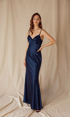 A woman stands against a beige backdrop wearing the Eureka 9711 Cowl-Neck Long Satin Prom Dress in navy blue, featuring thin straps and a draped neckline.