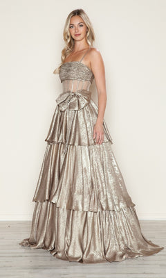A woman models the Long Prom Dress: Poly USA 9718, featuring metallic fabric, a tiered A-line skirt, thin straps, visible boning on the fitted bodice, and a large waist bow. She stands on light wood flooring against a neutral background.