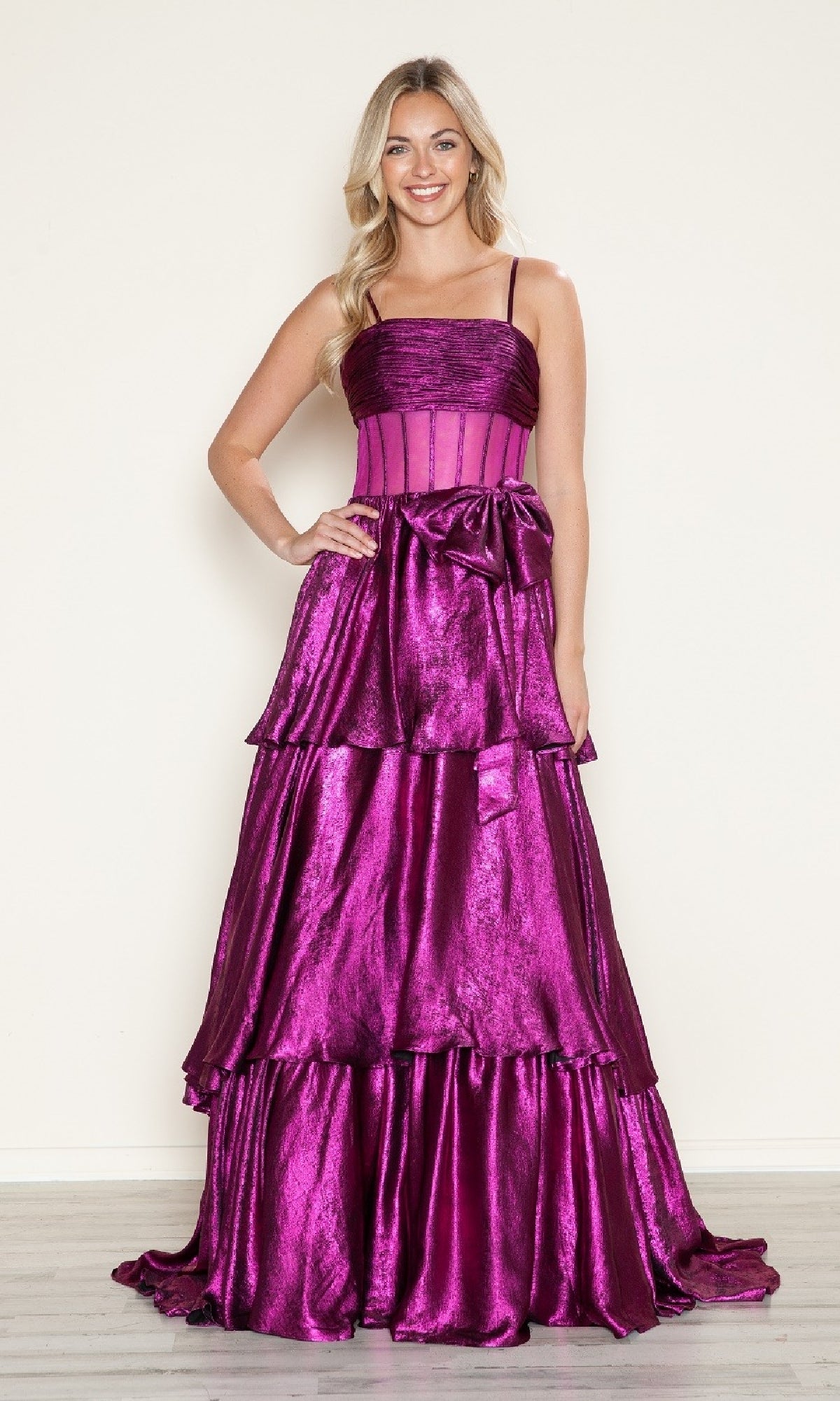 A woman smiles in the Long Prom Dress: Poly USA 9718, a floor-length, shiny magenta metallic gown with tiered ruffles, thin straps, and a large bow at the waist.