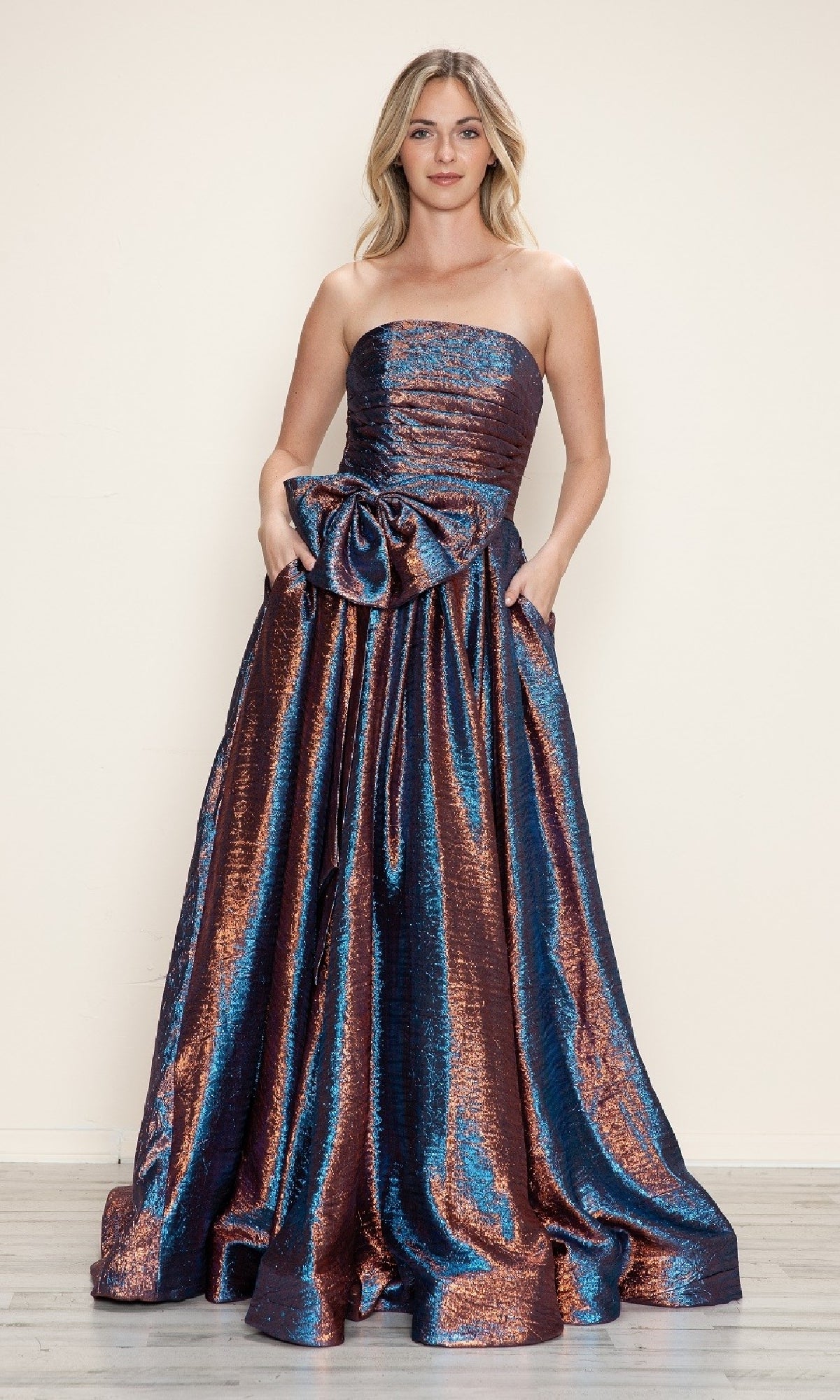 A woman models the Poly USA 9720 Side-Bow Strapless Prom Gown in metallic blue and copper, featuring a large waist bow, against a simple light background.