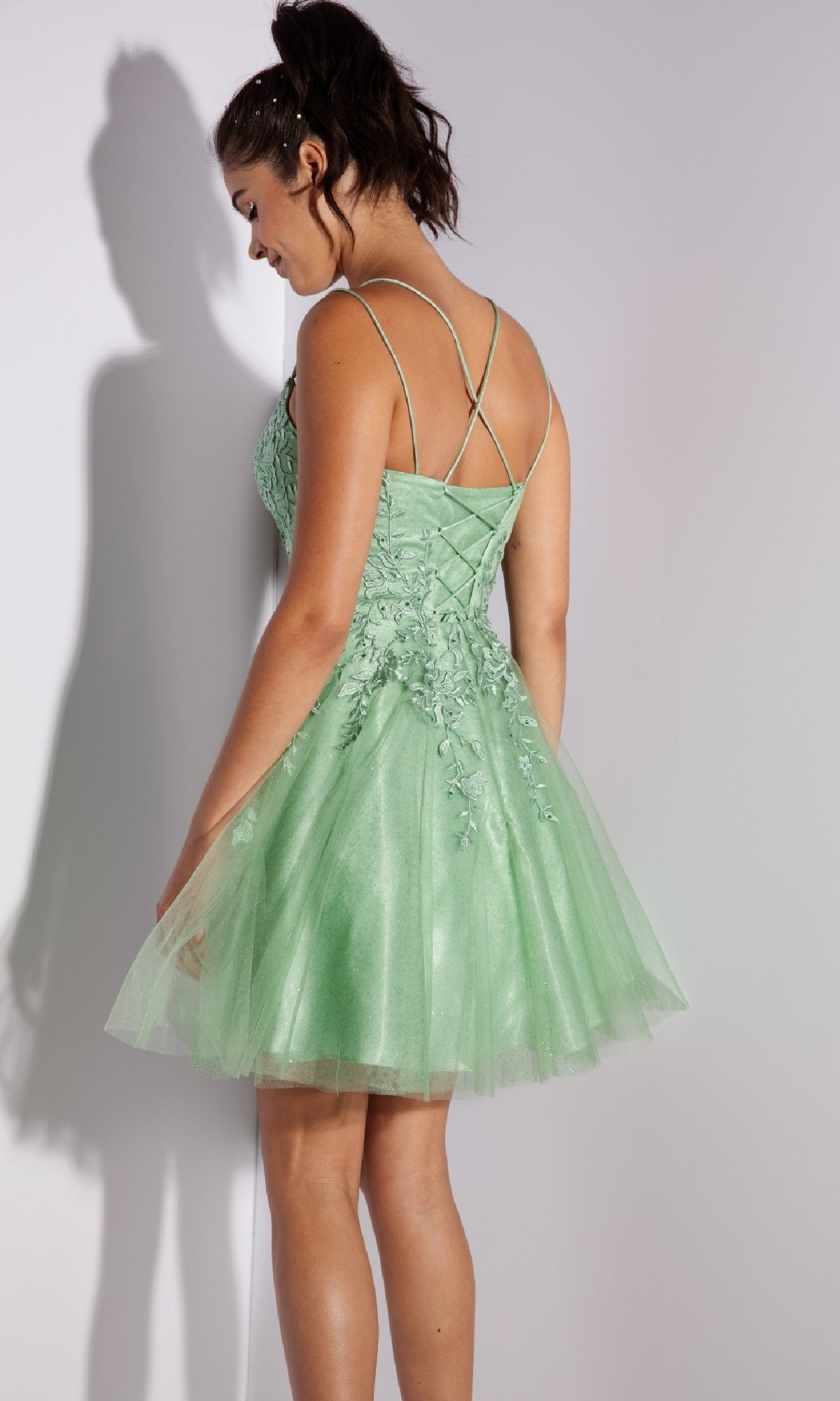 Woman wearing the Eureka 9727 Lace-Up Short Prom Dress in light green, a sleeveless tulle design with floral embroidery and crisscross back straps, stands looking down.