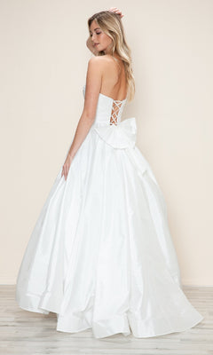 A woman wearing the Long Prom Dress: Poly USA 9754, featuring a strapless ruched bodice, adjustable lace-up back, and large bow, stands on a light wood floor with a plain beige background.
