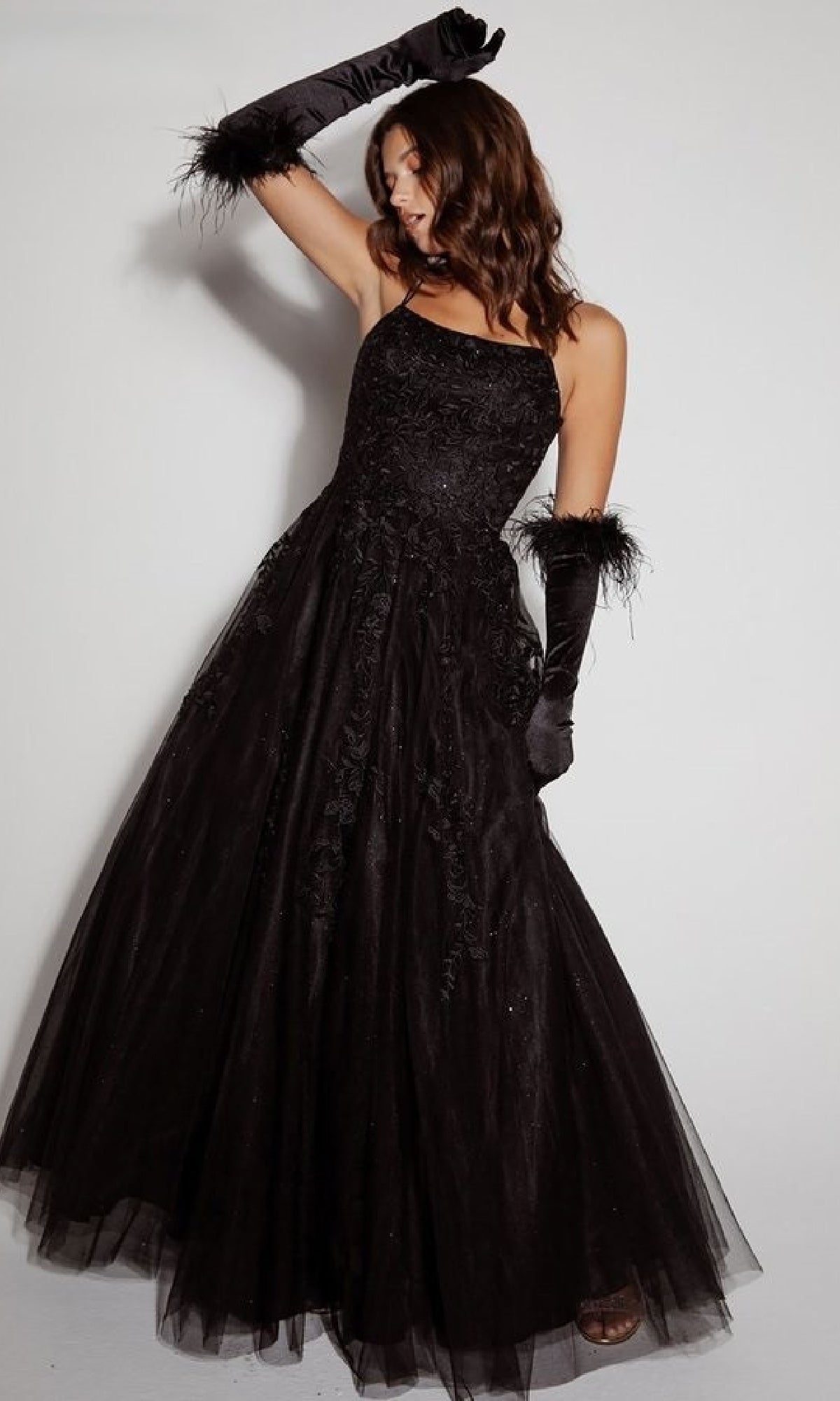 A woman models the Eureka CC0240 Lace-Up Long Lace Prom Ball Gown, featuring elegant lace details and a flattering silhouette, paired with feathered gloves, against a plain white background.