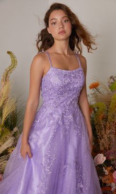A young woman stands before flowers and greenery, wearing the Eureka CC0240 Lace-Up Long Lace Prom Ball Gown—lavender, glittery, embroidered lace with a graceful silhouette.