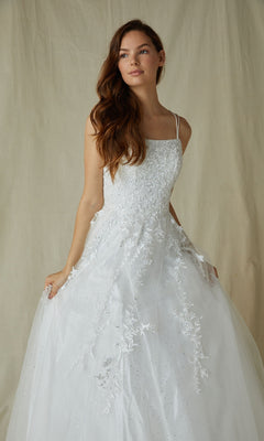 A woman in the Eureka CC0240 Lace-Up Long Lace Prom Ball Gown, featuring white floral embroidery, a tulle skirt, and lace-up back, poses against a beige fabric backdrop.
