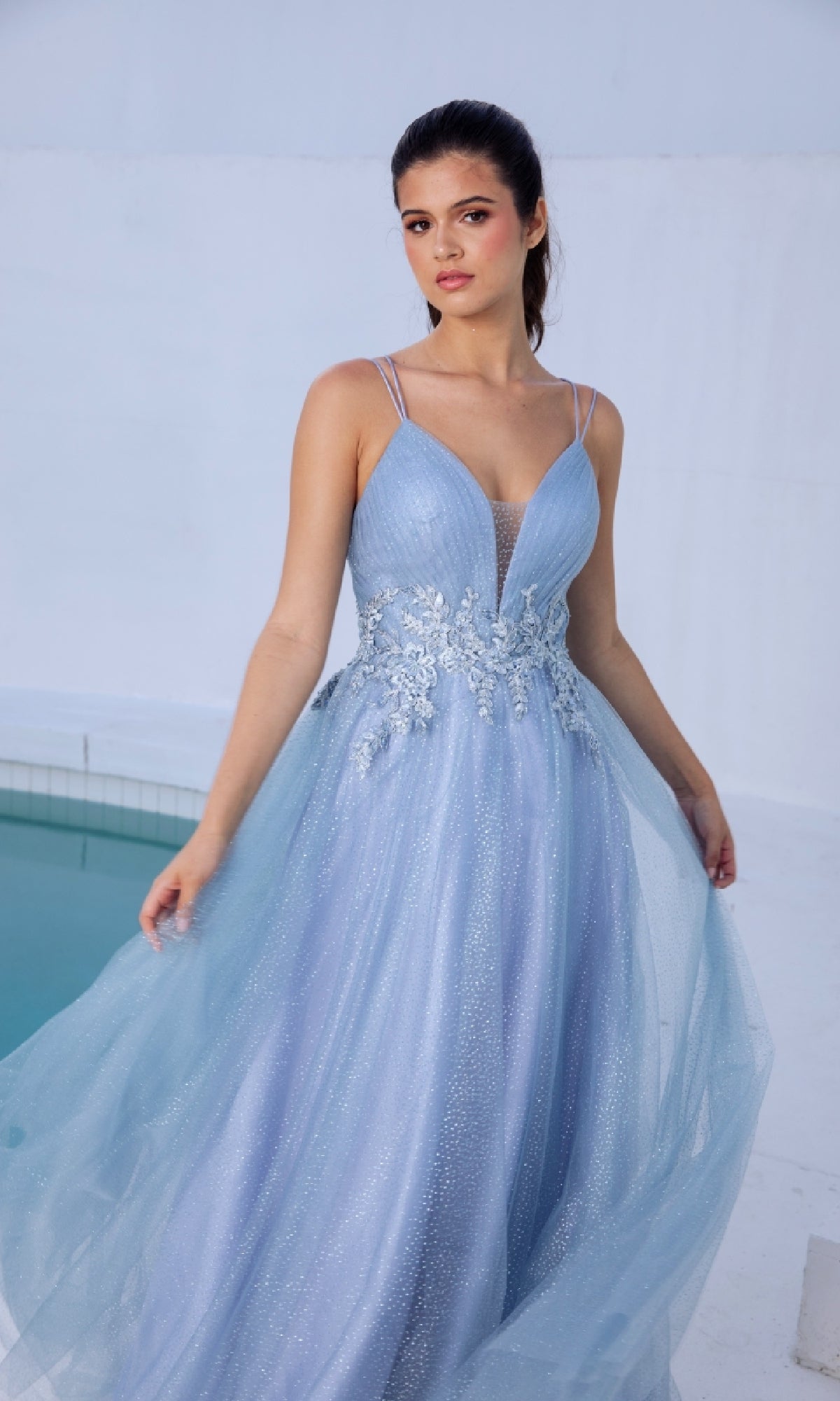 A woman wears the Lace-Up Long Shimmer Prom Dress: Eureka 9797, a light blue sleeveless gown with floral embroidery and a sparkly tulle skirt, standing beside a swimming pool against a white background.