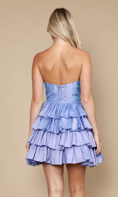 A woman with blonde hair models the Poly USA 9800 Ruffled Short Strapless Hoco Dress in iridescent lilac, featuring multi-layered ruffles as she stands against a beige background, facing away from the camera.
