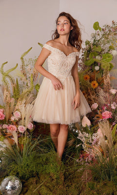 A young woman wears the Off-the-Shoulder Short Hoco Dress: Eureka 9833, standing indoors amid vibrant flowers and greenery.