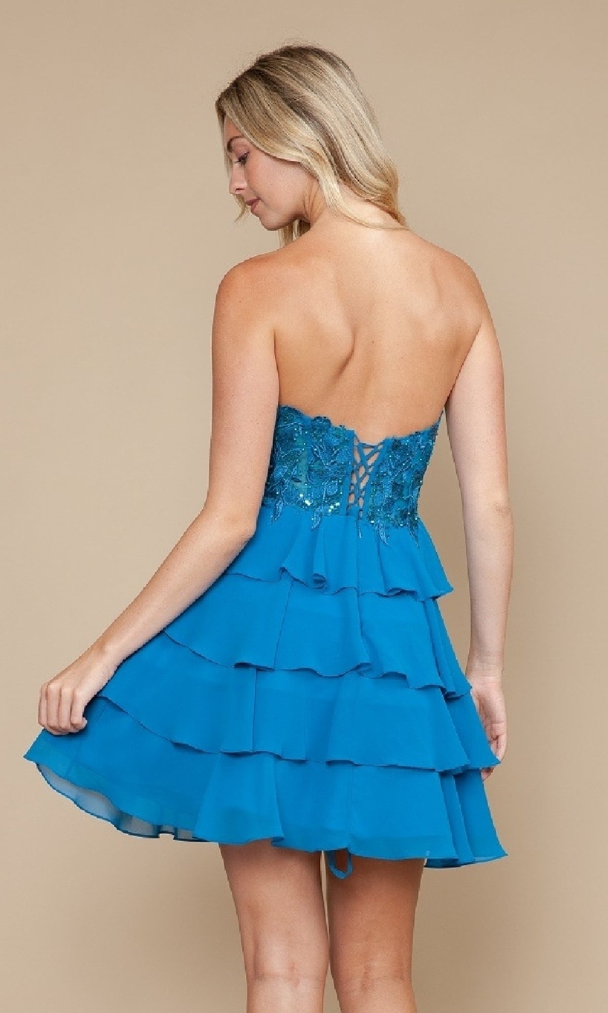 A woman wears the Strapless Short Ruffled Hoco Dress: Poly USA 9838, featuring a corset-style bodice, blue tiered ruffles, and floral embroidery, as she stands against a beige background.