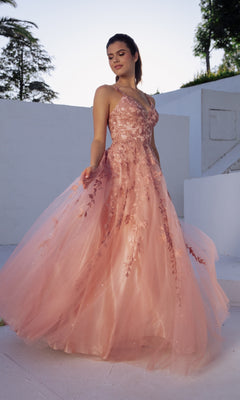 A woman stands outdoors on a white surface, wearing the Embroidered-Lace Long Prom Dress: Eureka 9858—a pink, sleeveless, floor-length formal dress with floral lace details—while looking slightly to the side.