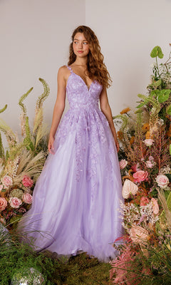 A woman in the Eureka 9858 Embroidered-Lace Long Prom Dress with floral appliqué stands among greenery and pink flowers.