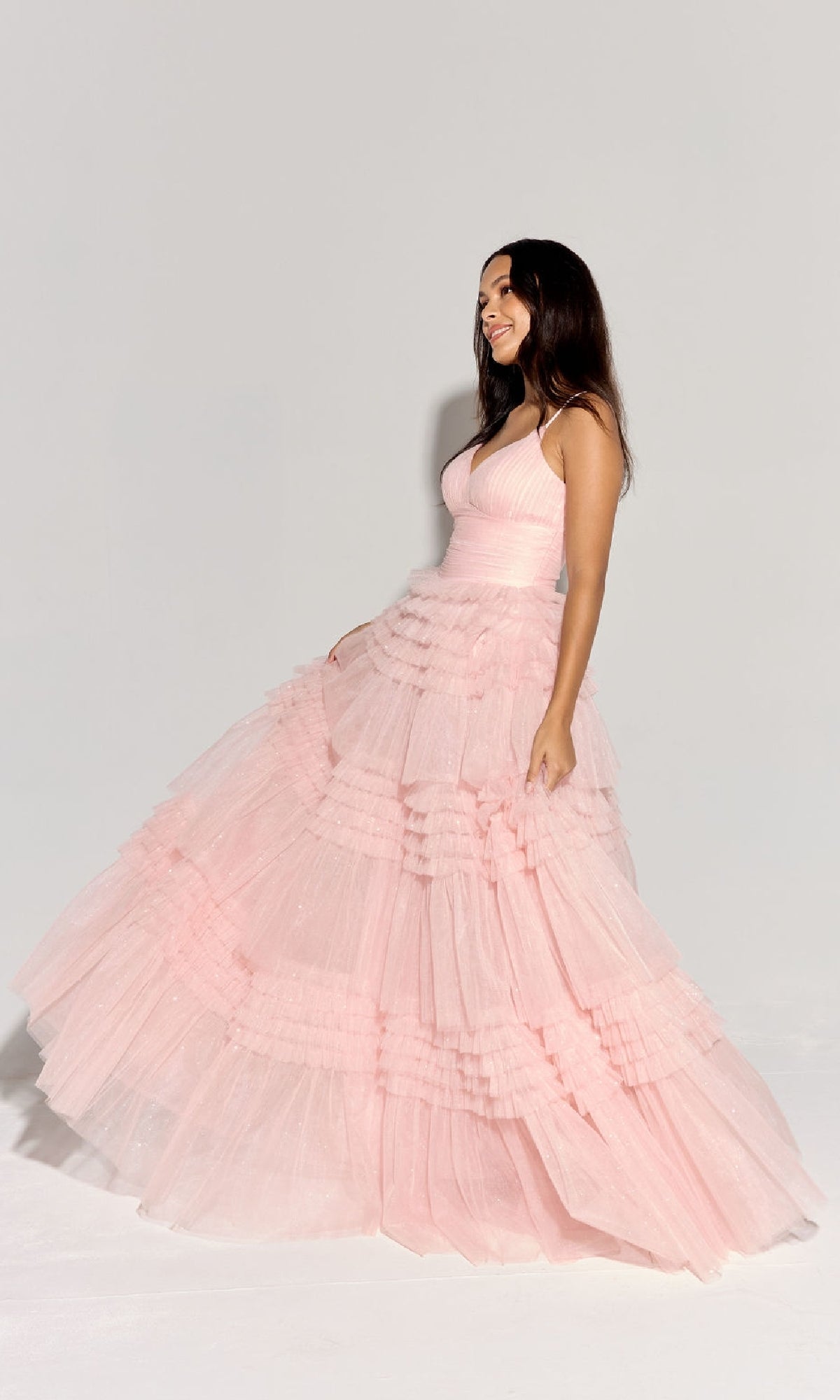 A woman stands sideways, smiling, wearing the Eureka 9888 Lace-Up Long Glitter-Tulle Prom Dress in light pink with thin straps and a lace-up back, against a plain light background.