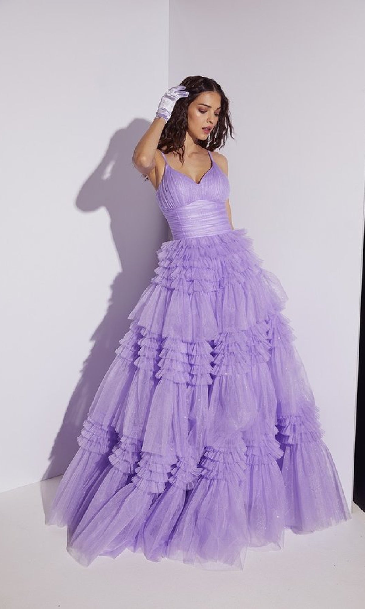 A woman models the Lace-Up Long Glitter-Tulle Prom Dress: Eureka 9888, featuring voluminous lavender tulle, layered ruffles, a sparkling finish, and lace-up back detail as she poses against a white wall with one gloved hand near her head.