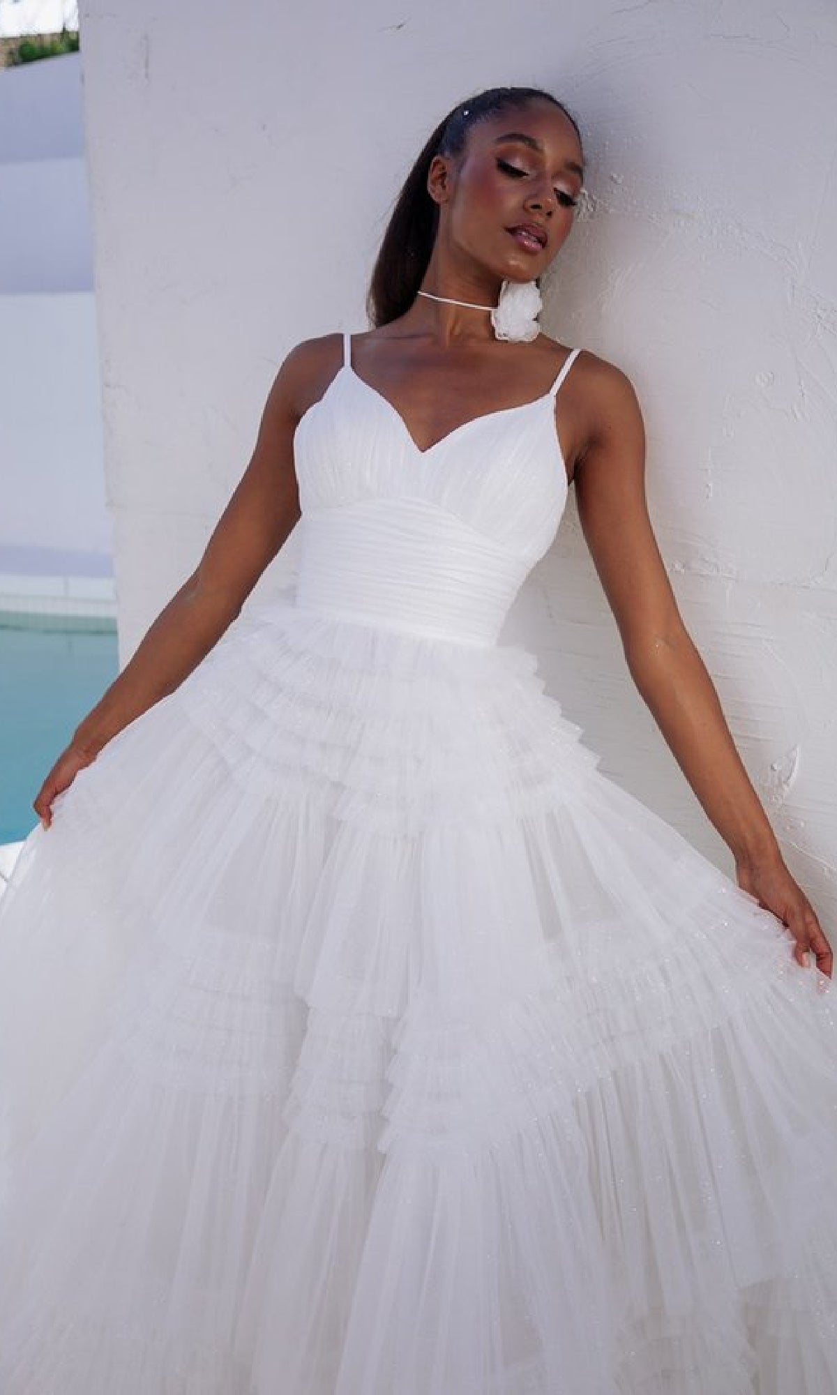 A woman models the Lace-Up Long Glitter-Tulle Prom Dress: Eureka 9888, featuring thin straps and a lace-up back, as she poses against a white textured wall holding out her skirt.