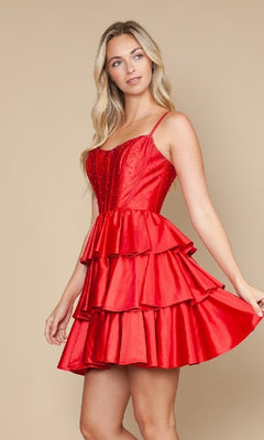 A woman wears the Lace-Up Short Ruffle Hoco Dress: Poly USA 9896, a sleeveless red fit-and-flare cocktail dress featuring a tiered ruffle skirt and fitted bodice, standing against a plain beige background.