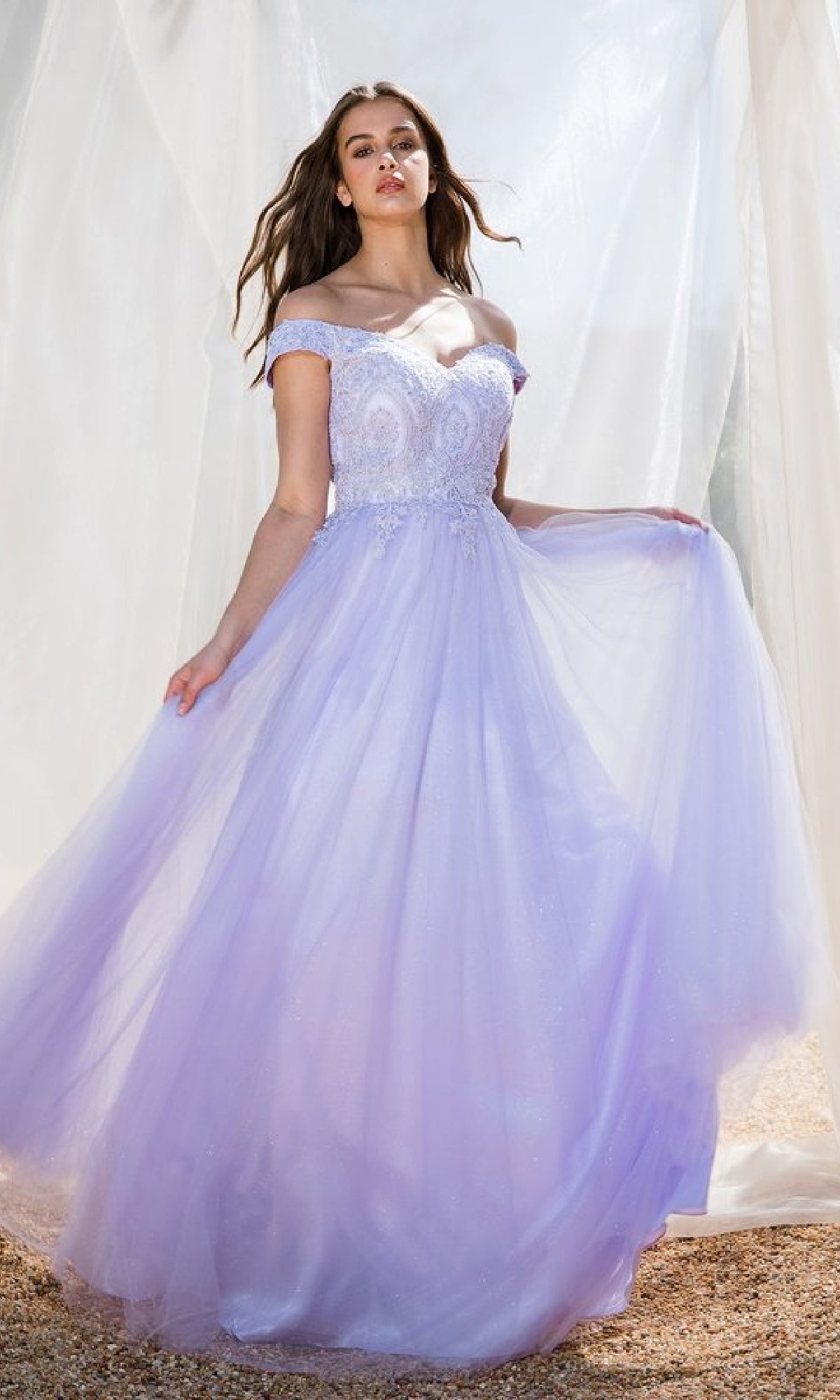 A woman outdoors wears the Off-Shoulder Glitter Prom Ball Gown Eureka 9899, a light purple dress with lace and tulle details, holding one side of the skirt.
