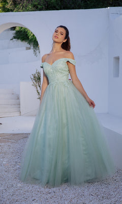 A woman poses outdoors before a striking white architectural backdrop, wearing the Eureka 9899 Off-Shoulder Glitter Prom Ball Gown with a fitted bodice and tulle skirt in light green.