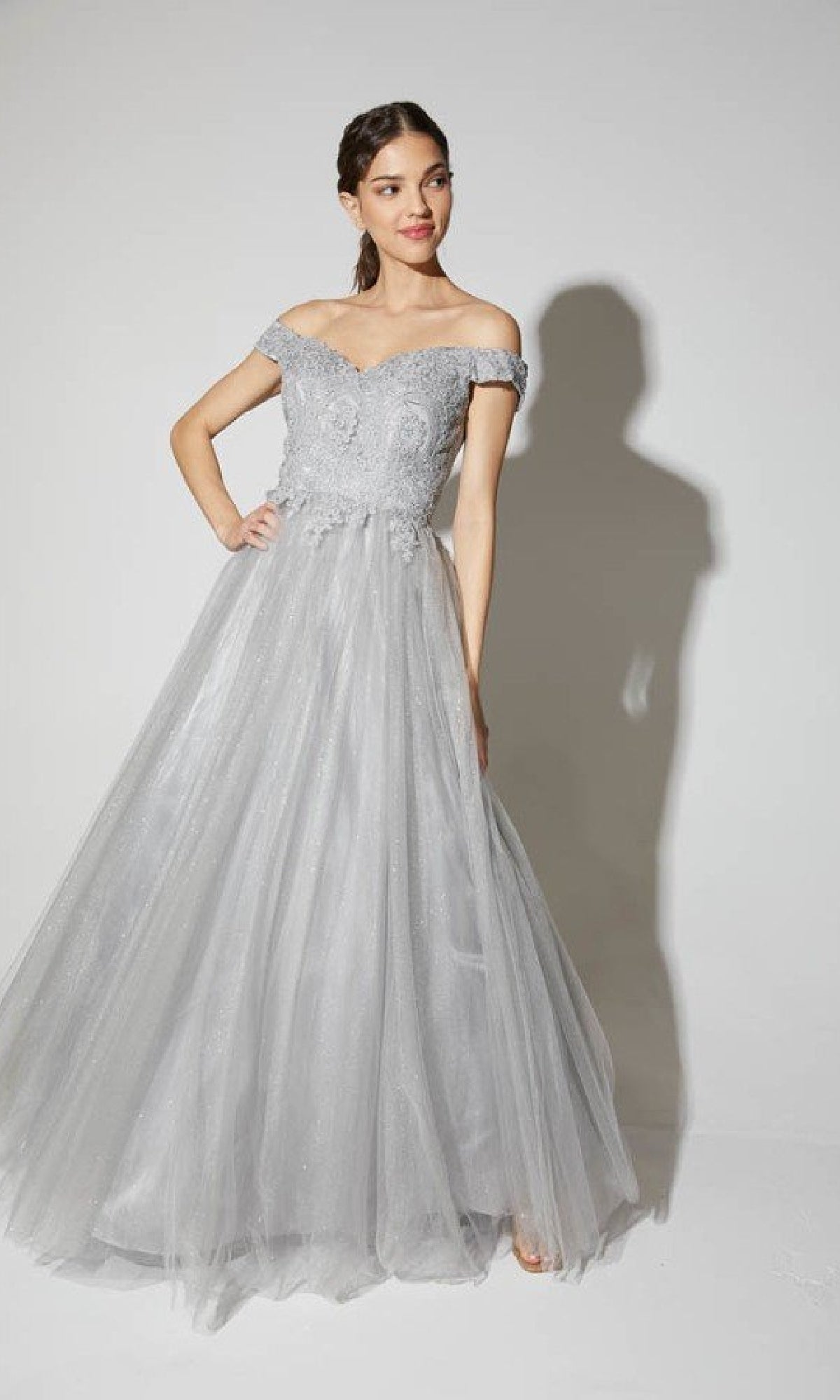 A woman poses against a plain white background in the Off-Shoulder Glitter Prom Ball Gown: Eureka 9899, featuring a light gray tulle skirt, shimmering glitter details, an off-the-shoulder neckline, and a lace bodice.