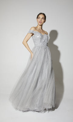 A woman poses in the Off-Shoulder Glitter Prom Ball Gown: Eureka 9899, a light gray dress with a fitted embellished bodice and full glitter-tulle skirt, creating an elegant look against a plain white background.