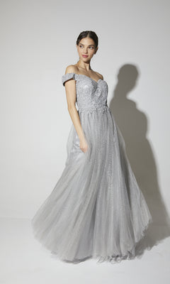 A woman poses in the Off-Shoulder Glitter Prom Ball Gown: Eureka 9899, featuring a light gray, off-the-shoulder design with lace details and a tulle skirt, set against a plain white background.