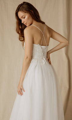 A woman in the Eureka 9901 Lace-Up Long Glitter Prom Ball Gown stands with her back to the camera, showcasing its crisscross straps and lace-up bodice against a neutral fabric backdrop.
