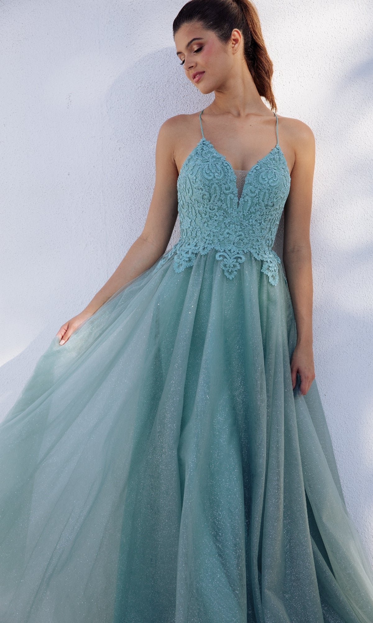 A woman in the Lace-Up Long Glitter Prom Ball Gown Eureka 9901, a sleeveless light blue lace and tulle evening dress, stands against a white wall holding one side of her gown.