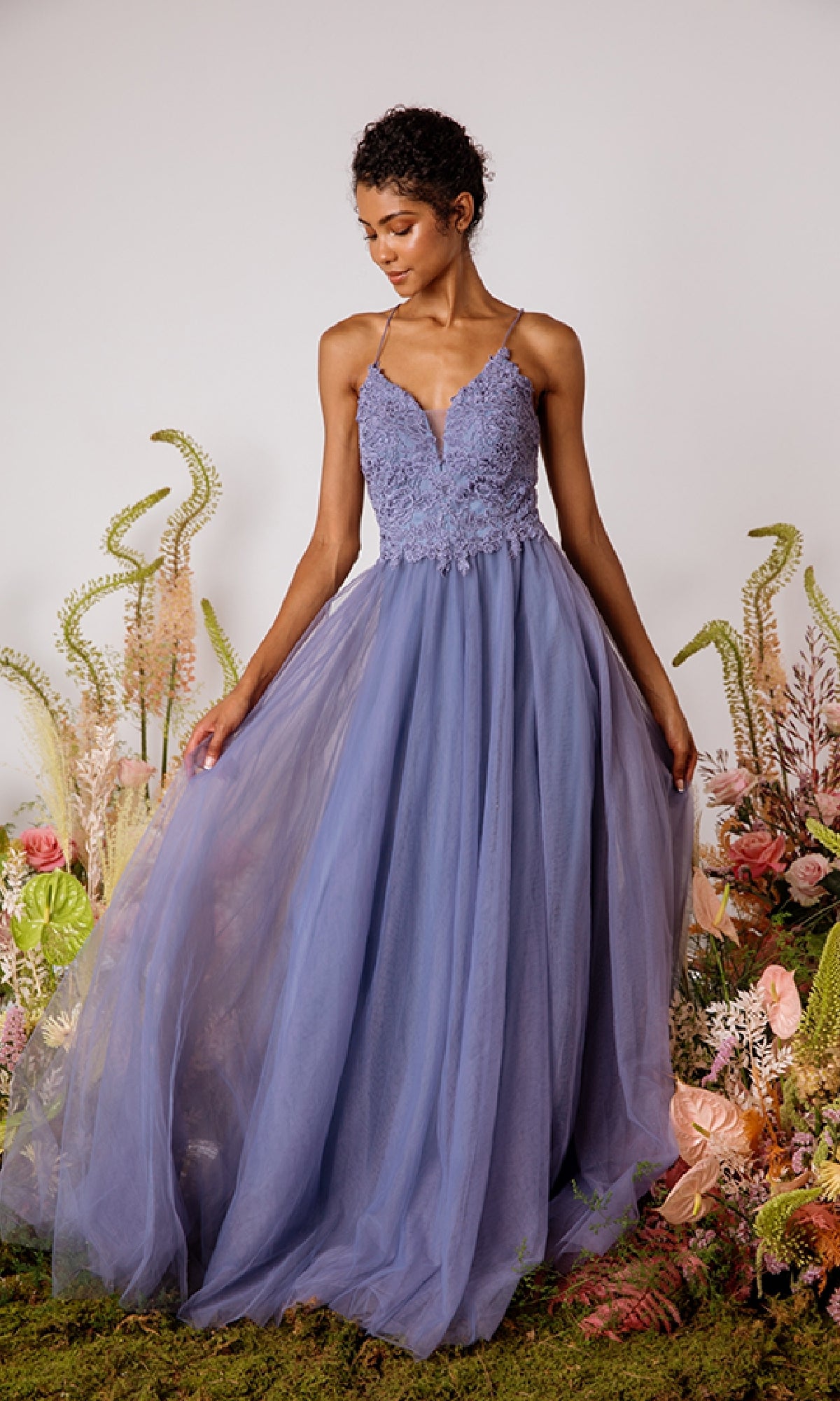 A woman wearing the Lace-Bodice Long Glitter Prom Dress Eureka 9902 with lavender lace, tulle skirt, and spaghetti straps stands amid assorted flowers and greenery.
