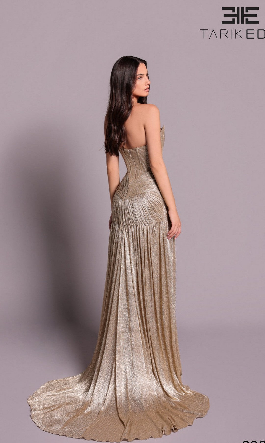 A woman with long brown hair wears the Tarik Ediz 99055 long formal dress in metallic gold, featuring a strapless design and flowing train, standing against a plain light purple background.