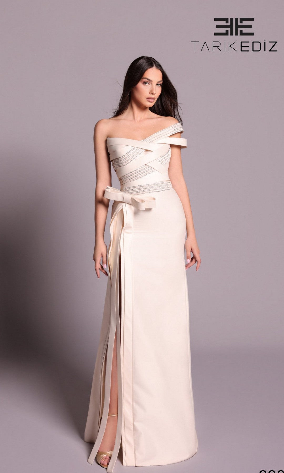 A woman wears the Tarik Ediz 99093 long formal dress in vanilla, featuring off-shoulder design, ribbon details, and a front slit, standing against a plain background with the Tarik Ediz logo.