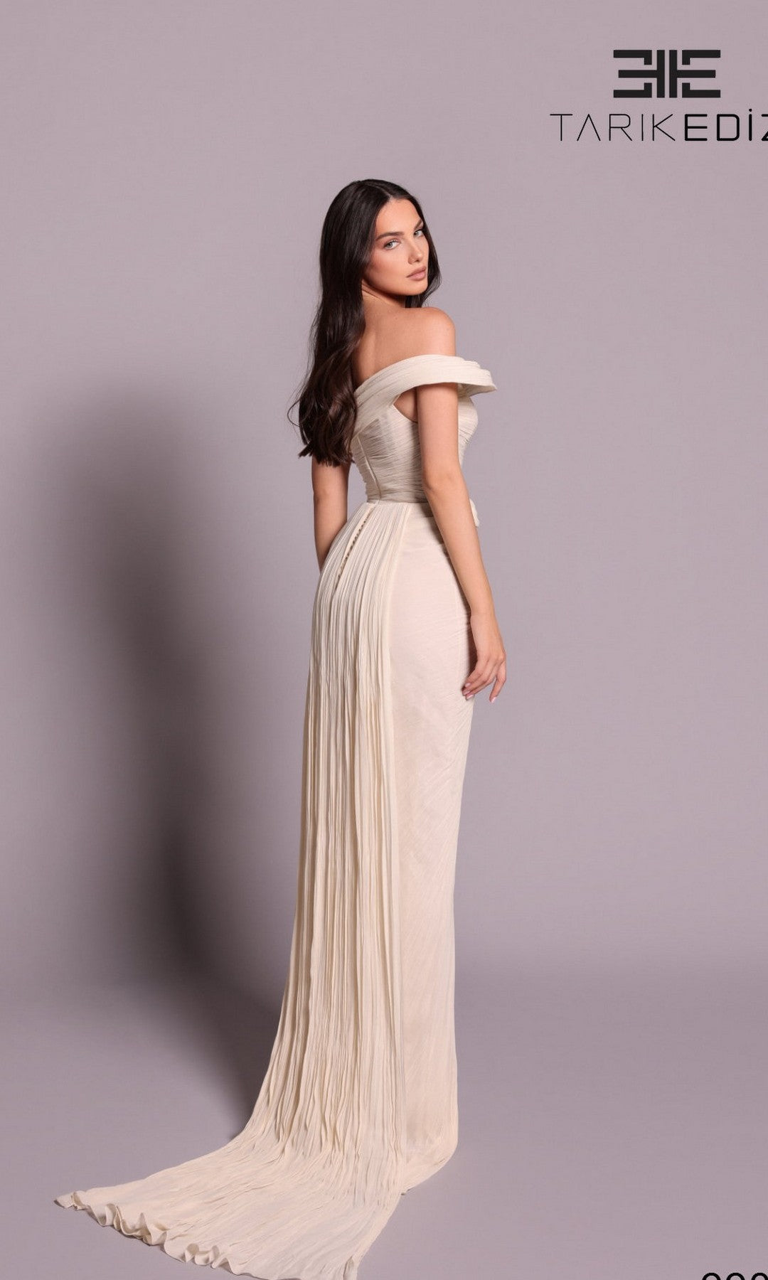 A woman poses against a plain light background in the Tarik Ediz 99095 Long Formal Dress, a cream-colored, off-shoulder, floor-length gown, with her body turned slightly to the side.