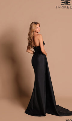 A woman wears the Tarik Ediz 99096 long formal dress, standing turned to the side against a beige background. The strapless black gown has a dramatic, flowing train perfect for special occasions.