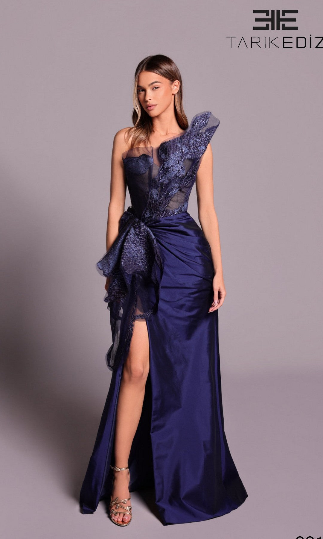 A woman wears the Tarik Ediz 99102 long formal dress in navy, featuring a one-shoulder design, asymmetrical lace bodice, high slit, and draped fabric details.