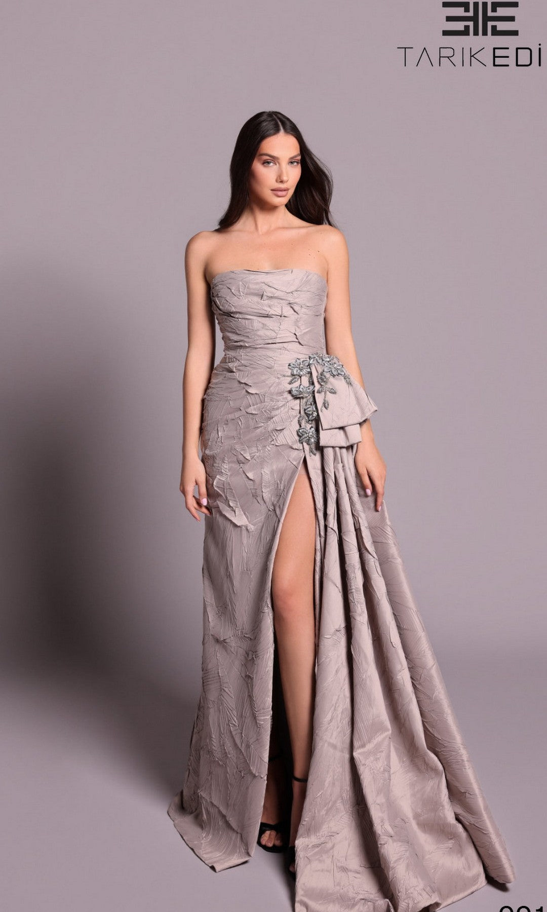 A woman stands against a plain backdrop wearing the Tarik Ediz 99105 long formal dress in mink—a strapless, textured gown with a thigh-high slit and floral embellishments. The Tarik Ediz logo appears in the top right corner.