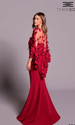 A woman wears the Tarik Ediz 99107 long formal dress, a fitted red couture gown with a mermaid silhouette and sheer overlay embellished with 3D floral appliqués, standing against a plain backdrop.