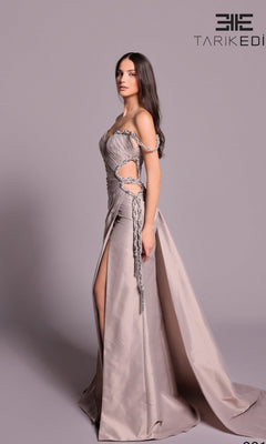 A woman stands in profile wearing the Tarik Ediz 99108 long formal dress in mink, featuring an off-shoulder sweetheart neckline, side cutouts, draping, and a long train. The background is plain light gray.