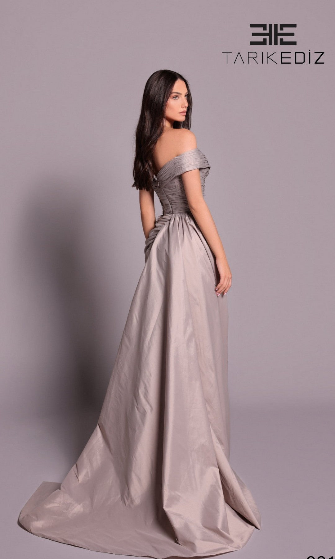 A woman poses sideways in the Tarik Ediz 99108, a silver-grey, off-the-shoulder, floor-length formal dress featuring a high-leg front slit. The 