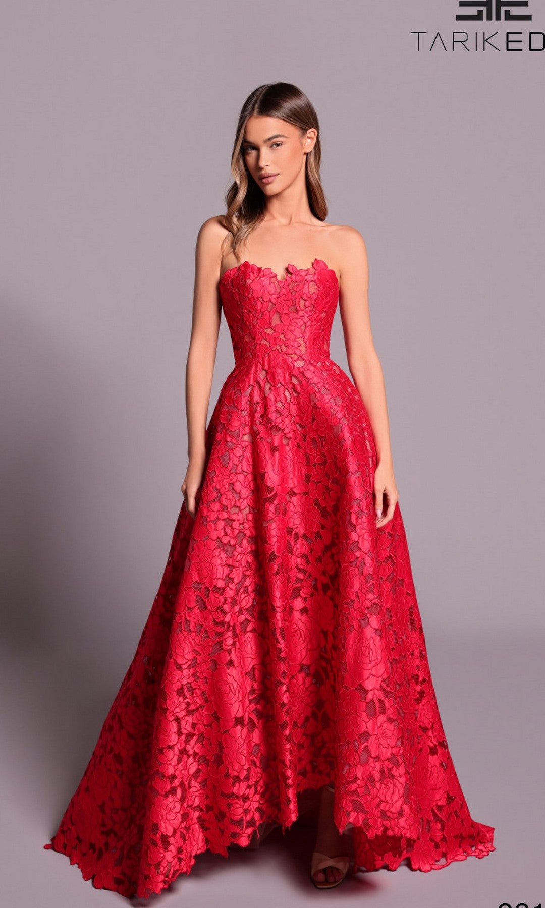 A woman stands against a plain background, wearing the Tarik Ediz 99111 long formal dress—a strapless, floor-length red gown with intricate floral lace detailing.
