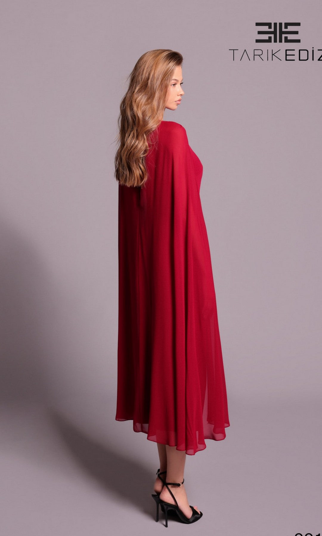 A woman with long wavy hair models the Tarik Ediz 99115, a flowing ankle-length red formal dress with cape sleeves and black heels, standing sideways against a plain light purple background.