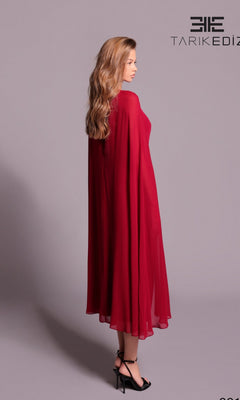A woman with long wavy hair models the Tarik Ediz 99115, a flowing ankle-length red formal dress with cape sleeves and black heels, standing sideways against a plain light purple background.