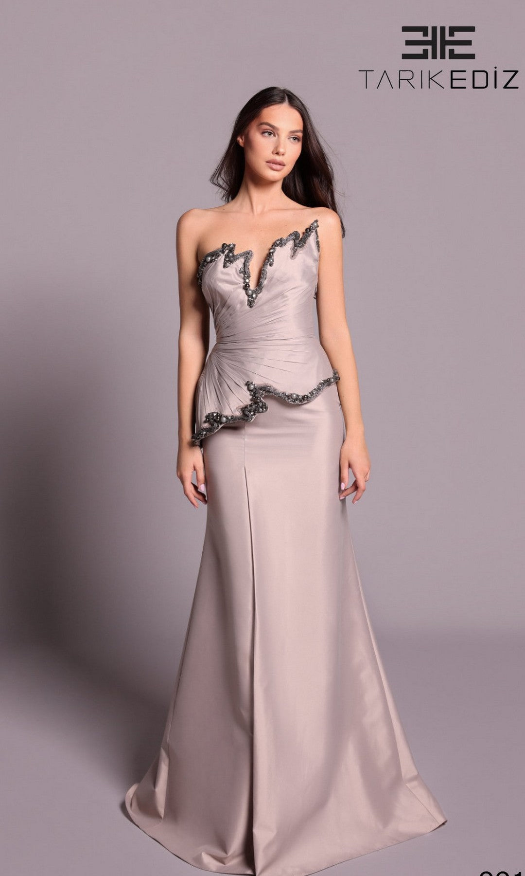 A woman models the Tarik Ediz 99119 long formal dress in mink, a strapless taffeta gown with black trim and peplum detail, standing before a light gray background with the brand’s logo visible.