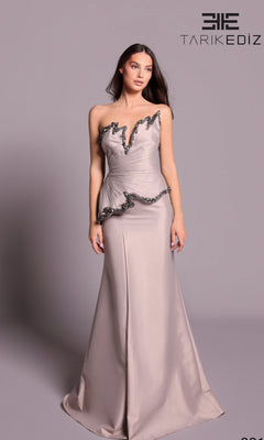 A woman models the Tarik Ediz 99119 long formal dress in mink, a strapless taffeta gown with black trim and peplum detail, standing before a light gray background with the brand’s logo visible.