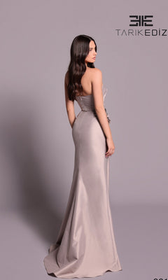 A woman in the Tarik Ediz 99126 long formal dress, strapless and fitted, stands facing away against a plain background, highlighting the gown's elegant floor-length design with the Tarik Ediz logo at the top right.