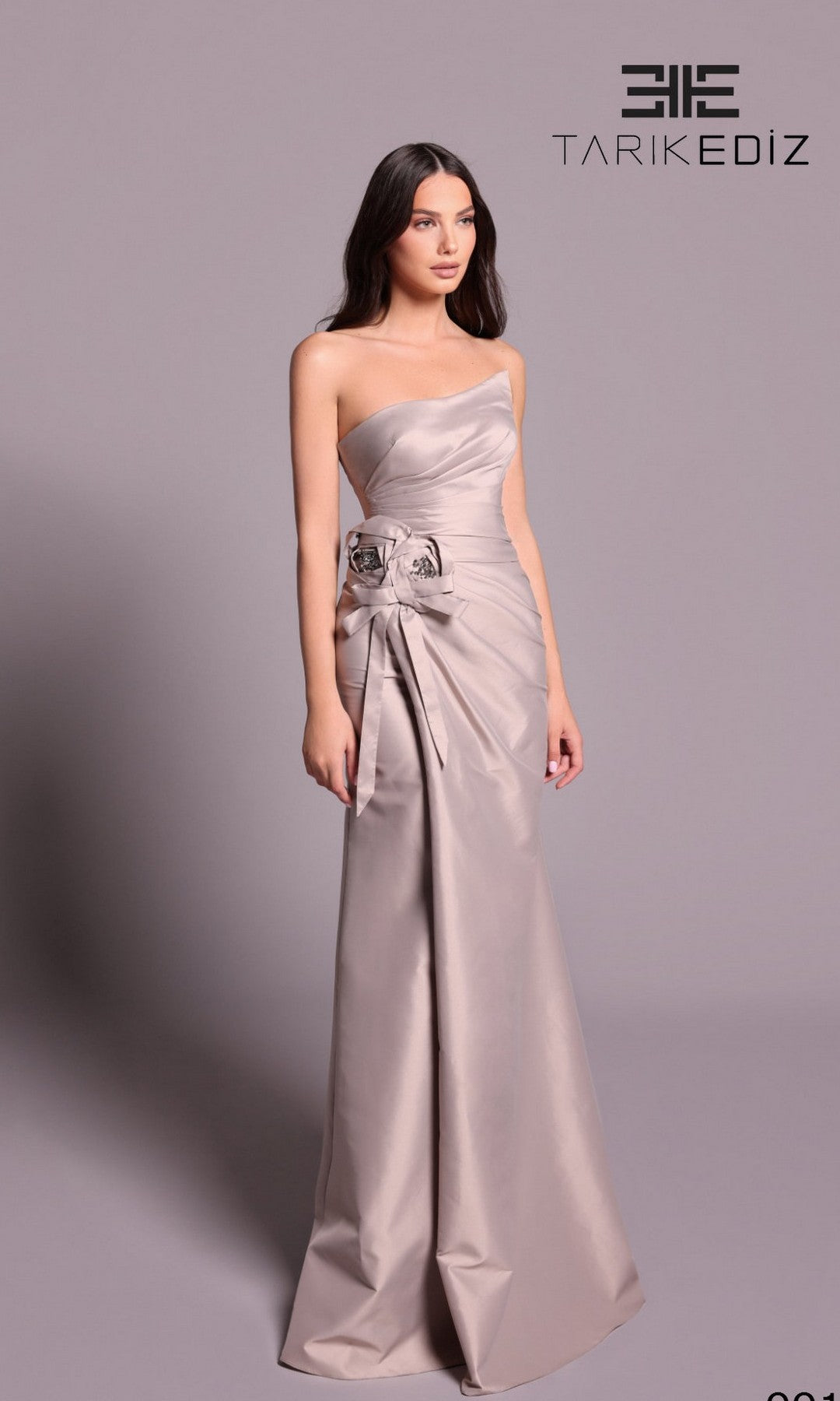 A woman models the Tarik Ediz 99126 long formal dress in mink, a strapless, floor-length gown with side floral detail. She poses against a plain backdrop with the 