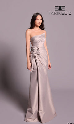 A woman models the Tarik Ediz 99126 long formal dress in mink, a strapless, floor-length gown with side floral detail. She poses against a plain backdrop with the "TARIK EDIZ" logo displayed in the top right corner.